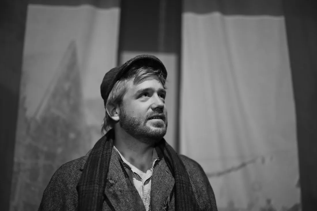 Ben Tissell as Toby Veck, in cap and scarf, looking up toward the bell tower.